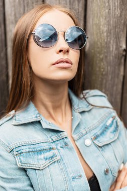 Şık güneş gözlüklü, güzel bir mankenin moda portresi ahşap bir duvarın yanında duruyor.