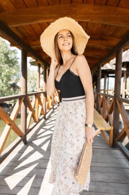 Yazın, şık bir hasır şapkayla, siyah stil dantelli bir bluzla kumsalda, ahşap terasın yanında yürüyen, mutlu, beyaz bir kadın.