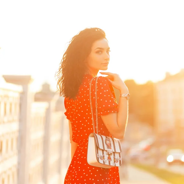 美しい若いです巻き女性でヴィンテージ赤ドレスでファッションハンドバッグはオレンジの夕日で通りを歩くとカメラを見て — ストック写真