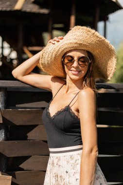 Moda yaz gözlüklü, örülmüş hasır şapkalı, ahşap çitin yanında dinlenen ve tatilde dinlenen güzel beyaz bir kız.