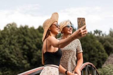 Gülümseyen güzel bir genç kadın ve güneş gözlüklü yaşlı mutlu bir anne şık yaz kıyafetleri içinde ortak bir selfie çektirip güneşli bir hafta sonunda dinleniyorlar.