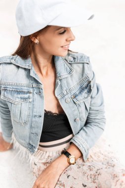 Kot ceketli, siyah dantelli, etekli, mavi şapkalı mutlu genç kadın kumsalda beyaz kumsalda oturuyor.