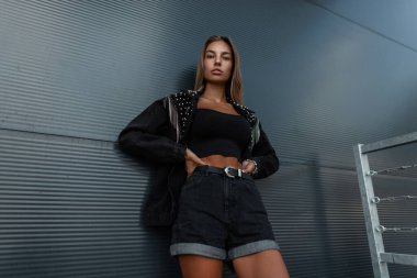 Siyah moda ceketli, kolsuz tişörtlü ve kot şortlu güzel beyaz kadın mankeni sokaktaki modern metal duvarın yanında duruyor. Kentsel kadın tarzı kıyafet