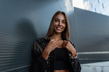 Güzel bir gülümsemesi olan, siyah tişörtlü şık kot ceketli mutlu genç bir kadın şehrin modern metal binasının yanında duruyor.