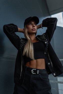 Siyah kot ceketli, kot şortlu ve kolsuz gömlekli havalı model kız şehirde koyu metal bir binanın yanında poz veriyor.