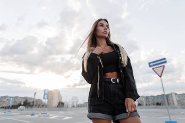 Siyah kot ceketli, siyah şortlu ve kolsuz tişörtlü güzel bir manken şehirde yürüyor.