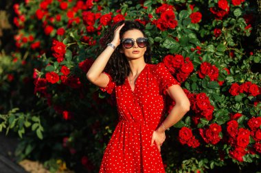 Güneş gözlüklü, kırmızı elbiseli, zarif, kıvırcık bir kadın gün batımında güllerin yanında bahçede poz veriyor.