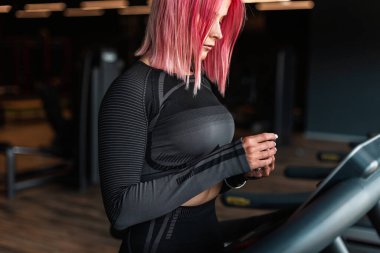 Siyah spor kıyafetleri içinde genç ve güzel bir bayan kulaklık takıyor ve spor salonundaki koşu bandında koşmaya hazırlanıyor.