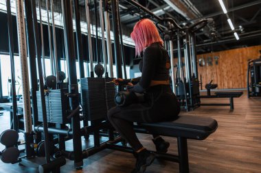 Güzel, modaya uygun, siyah spor giyimli, pembe saçlı atletik kız simülatörün yanında oturuyor ve spor salonunda egzersiz yapmaya hazırlanıyor.