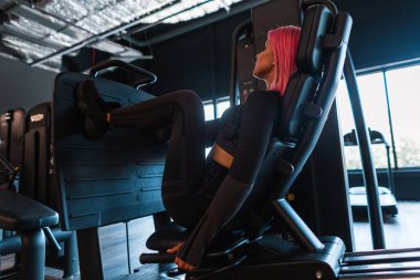 Siyah spor ayakkabılı, pembe saçlı güzel bir kız simülatöre oturur ve bacaklarını pompalar.