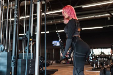 Siyah eşofmanlı, pembe saçlı güzel bir kadın spor salonundaki simülatöre kas yapar.