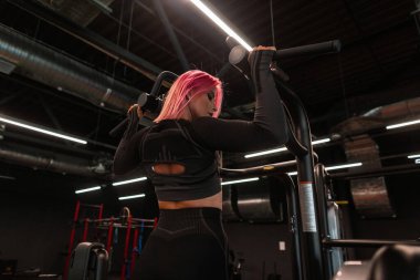 Pembe saçlı, şık siyah spor elbiseli güçlü genç bir kız koridorda kendini yukarı çekiyor.