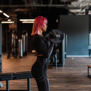 Spor kıyafetleri giyen sağlıklı spor kızı antrenman yapmaya başladı ve spor salonunda tabakları ve yan manzarası olan barı tuttu.