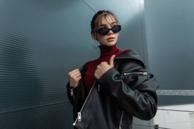 Modern şehrin metal duvarının yanında siyah kot ceket ve bordo süveter giymiş şık güneş gözlüklü, güzel, modaya uygun bir kadın modeli.