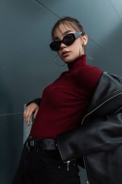 Moda gözlüklü, şık, siyah deri ceketli, bordo süveterli, şık, genç bir kadın sokakta metal bir duvarın yanında duruyor.