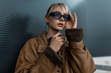 Şehirde siyah metal bir duvarın yanında şık güneş gözlüklü, klasik kahverengi deri ceketli güzel bir kadının moda portresi.