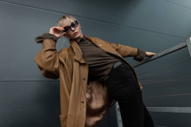 Moda gözlüklü, şık kahverengi deri ceketli ve metal duvarın yanında siyah kot giyen genç ve güzel bir kadın.