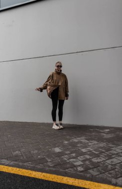 Şık, zarif, deri ceketli, siyah tozlu ve spor ayakkabılı genç bir kadın sokakta gri bir şehir duvarının yanında duruyor.