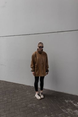 Şık, kahverengi ceketli, güneş gözlüklü, siyah kotlu genç model kız şehrin en az gri duvarının yanında duruyor.