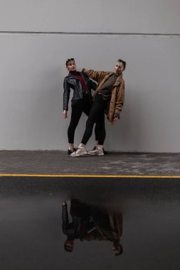 Şık deri ceketli ve siyah kotlu iki şık şehir modeli şehirde gri bir duvarın yanında poz veriyor.