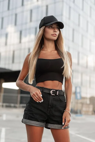 美しい若いモデルの女性は 街の魅力的なベルトの散歩と黒のキャップ 黒の空白のタンクトップとデニムのショートパンツを身に着けています — ストック写真
