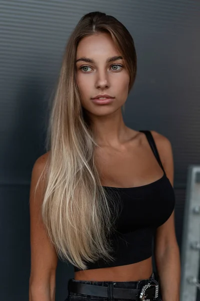 Portrait fashion of a pretty fashionable woman with a beautiful face with green eyes in a black stylish tank top and jeans stands near a metal wall outdoors