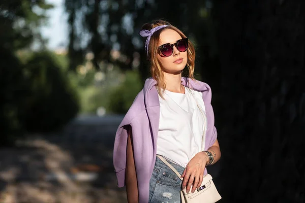 白いTシャツと紫のパーカーとジーンズのショートパンツでサングラスを持つ官能的な美しい若い女性の明るいファッションの肖像画 — ストック写真