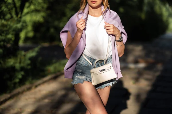ファッショナブルな女の子でリッピングデニムショートパンツにアクセサリーハンドバッグで紫のスウェットシャツ歩く路上 — ストック写真