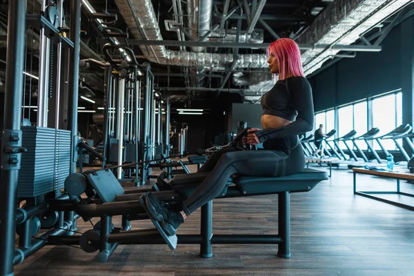 一个身材纤细 一头粉红头发 身穿黑色运动服的年轻貌美姑娘坐在模拟器旁 把她的背抽进体育馆 — 图库照片