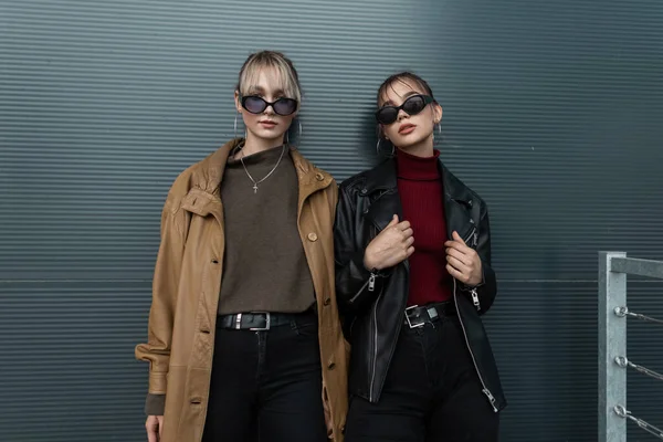 时尚双语 两位穿着时尚太阳镜 穿着皮夹克 穿着黑色牛仔裤的年轻女士站在街上的金属墙边 — 图库照片