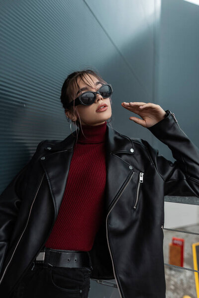 Крутая молодая модель женщина в старинных солнцезащитных очках в модном черном кожаном пиджаке с черными джинсами и бордовым свитером позирует возле металлического современного здания в городе