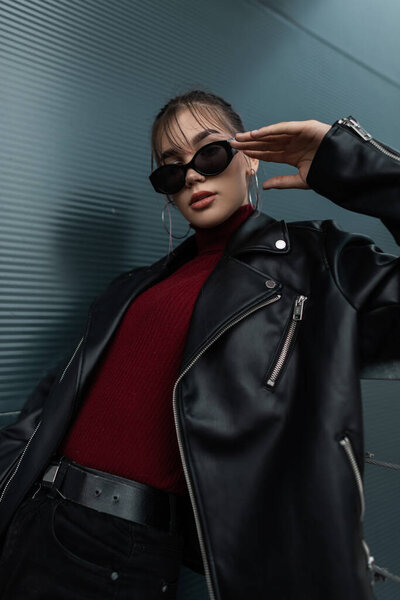 Крутая молодая модель женщина в модных солнцезащитных очках в стильной джинсовой куртке с бордовыми свитерами позирует возле металлической стены в городе