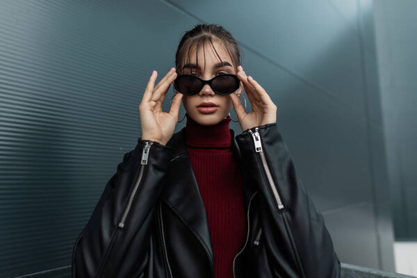 Довольно стильная девушка в черной кожаной куртке с молнией надевает прохладные солнцезащитные очки в городе. Осенняя коллекция весенней женской одежды