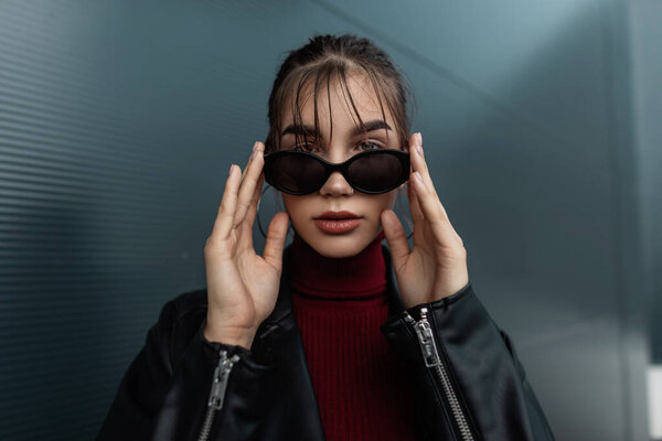 Стильный портрет красивой молодой женщины в модной черной кожаной куртке с молнией в бордовом свитере выпрямляет солнечные очки в городе, смотрит в камеру