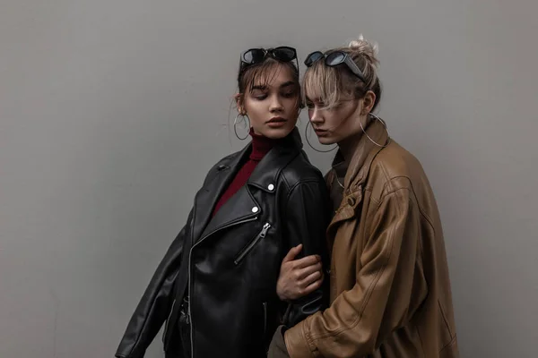在城市灰墙附近 两个漂亮的年轻时尚女模特戴着时尚太阳镜 穿着老式皮夹克 — 图库照片