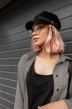 Moda mankeni, şapka takmış pembe saçlı, gözlüklü, şık bir kadın modeli şehirde gri bir duvarın yanında duruyor.
