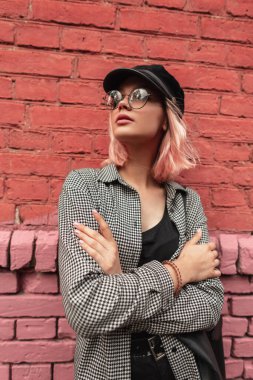 Modayı takip eden, gözlüklü, şapkalı, tişörtlü, sokakta duran kırmızı tuğlalı duvarın yanında poz veren şık bir kadın mankeni.