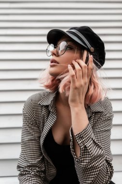 Havalı bir yüzü, zarif yuvarlak gözlükleri ve klasik bir günlük gömleği olan şapkası olan güzel bir genç kızın moda portresi ahşap bir duvarın yanında poz veriyor. Kadın portresi