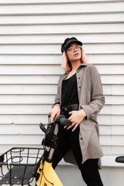 Gözlüklü, şapkalı, şık bir kadın. Siyah tişörtlü, kot pantolonlu, bisikletli, ahşap bir evin yanında gezen.