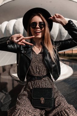 Güneş gözlüklü, klasik şapkalı, deri ceketli ve siyah çantalı komik genç kadın barış işareti yapıyor ve şehirde oturuyor.