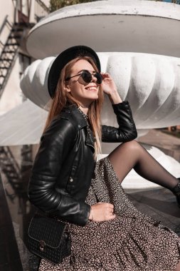 Güneş gözlüklü, şık deri ceketli ve siyah çantalı güzel, zarif, genç bir kız güneşli bir günde açık havada oturuyor.
