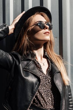 Güneşli bir günde, siyah gözlüklü, şık şapkalı, klasik elbiseli ve deri ceketli güzel bir kadının moda portresi.