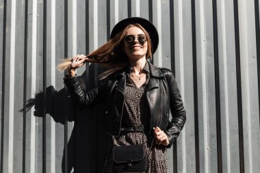 Moda kıyafetleri içinde, deri ceketli, şapkalı ve güneş gözlüklü neşeli genç kız güneşli bir günde metal bir arka planda duruyor.