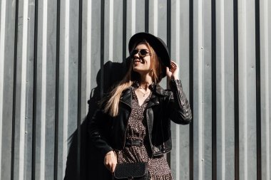 Güneş gözlüklü, siyah deri ceketli ve klasik elbiseli sevimli bir kız modeli güneşli bir günde metal bir duvarın yanında duruyor.