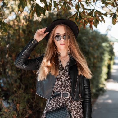 Güz güneşli kentte ağaçların yanında, modaya uygun siyah elbiseli, deri ceketli, güneş gözlüklü, şapkalı ve el çantalı güzel bir kadın model yürüyor.