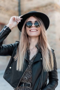 Güneşli bir günde, deri ceketli ve şık güneş gözlüklü güzel, mutlu bir kadının moda portresi.