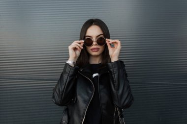 Siyah deri ceketli ve sweatshirtlü hippi bir kızın şık şehir portresi şehirdeki metal bir duvarın arka planında eski model güneş gözlüklerini düzeltiyor.