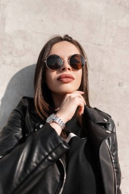 Gri beton arka planda, havalı siyah deri ceketli ve güneş gözlüklü güzel bir kızın moda portresi.
