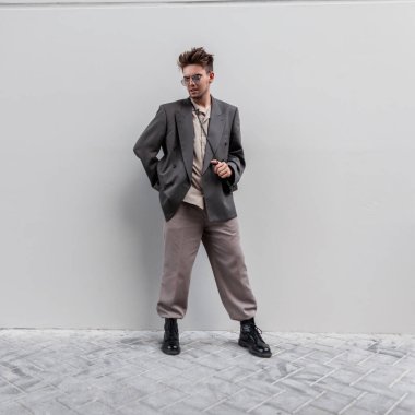 Tüylü, ceketli, güneş gözlüklü, gömlekli ve botlu şık bir genç adam şehirde gri bir duvarın yanında duruyor.