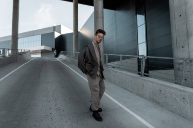 Moda mankeni, şık bir iş elbisesi içinde, spor ceketli ve gömlekli hippi bir adam şehirde modern bir binanın yanında yürüyor.